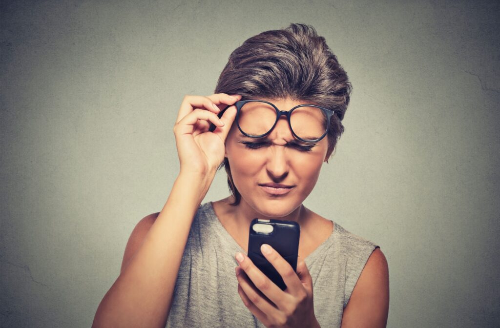 A picture of a younger individual slightly removing their glasses to squint and focus on their smartphone screen.