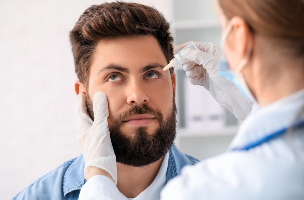 A person with a beard sits while an eye care professional gently administers eye drops during an exam.